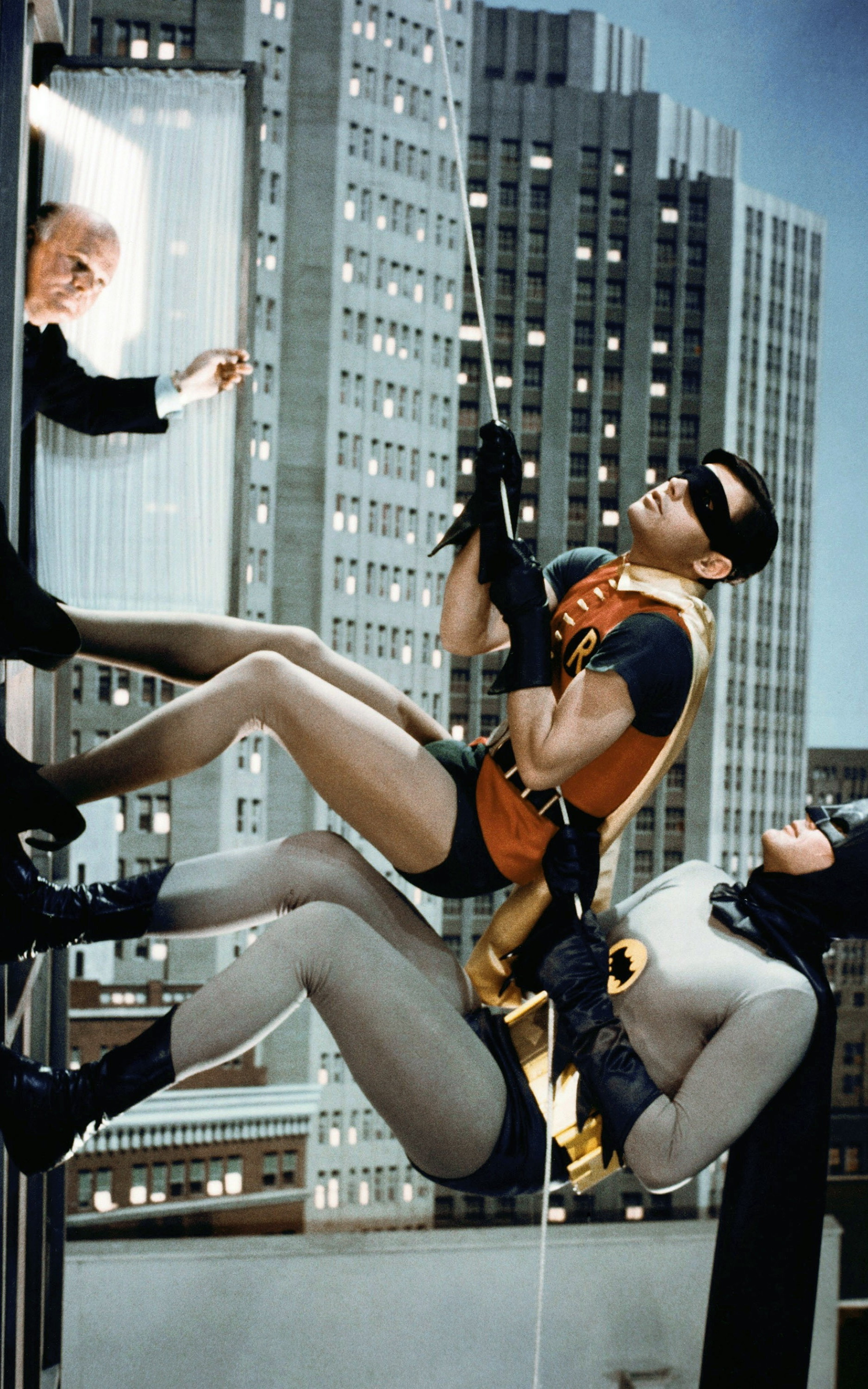 still from the 1960s Batman. Batman and Robin appear to be scaling a wall holding a thin rope. A man sticks his head out the window.