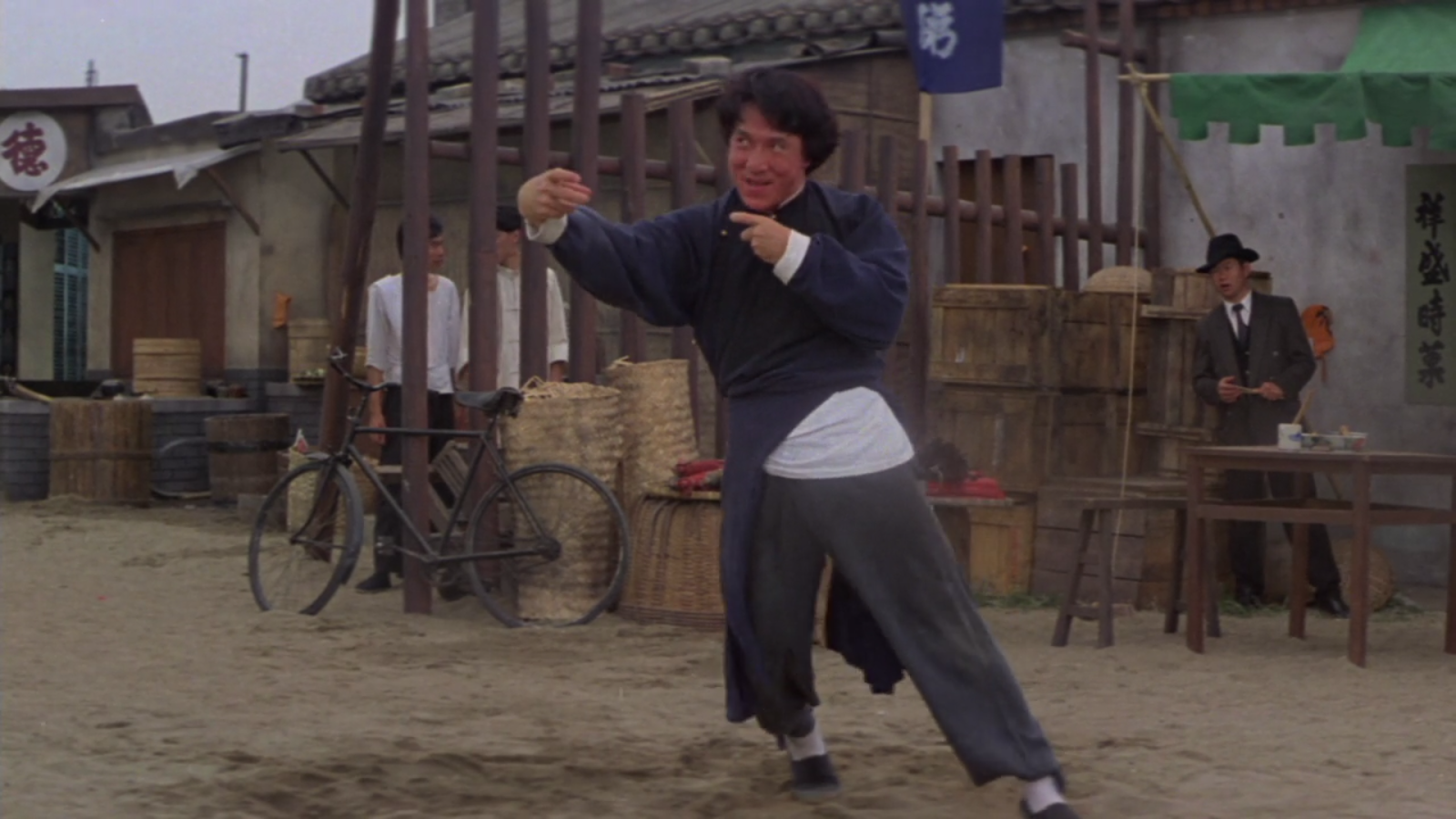A man (Jackie Chan) in early 20th century Chinese costume lists to the side as if drunk while holding his hand out in an awkward but aggressive manner. His face is red and he has a silly grin.
