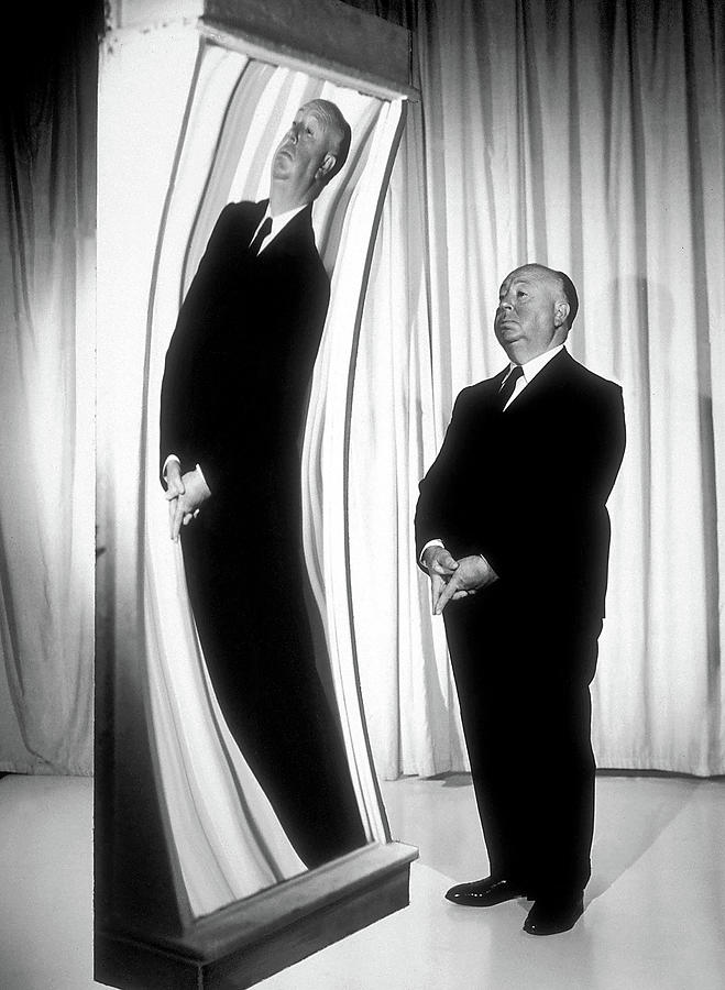 Black and white photo of Alfred Hitchcock standing in a suit in front of a funhouse mirror, his image in the mirror distorted to make him look tall and twisted.