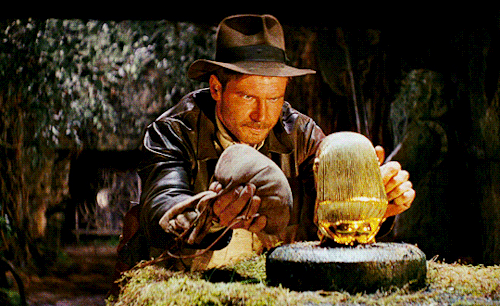 A man (Harrison Ford playing Indiana Jones) in a leather coat and dirty fedora prepares to replace a golden idol on an altar with a bag of sand.