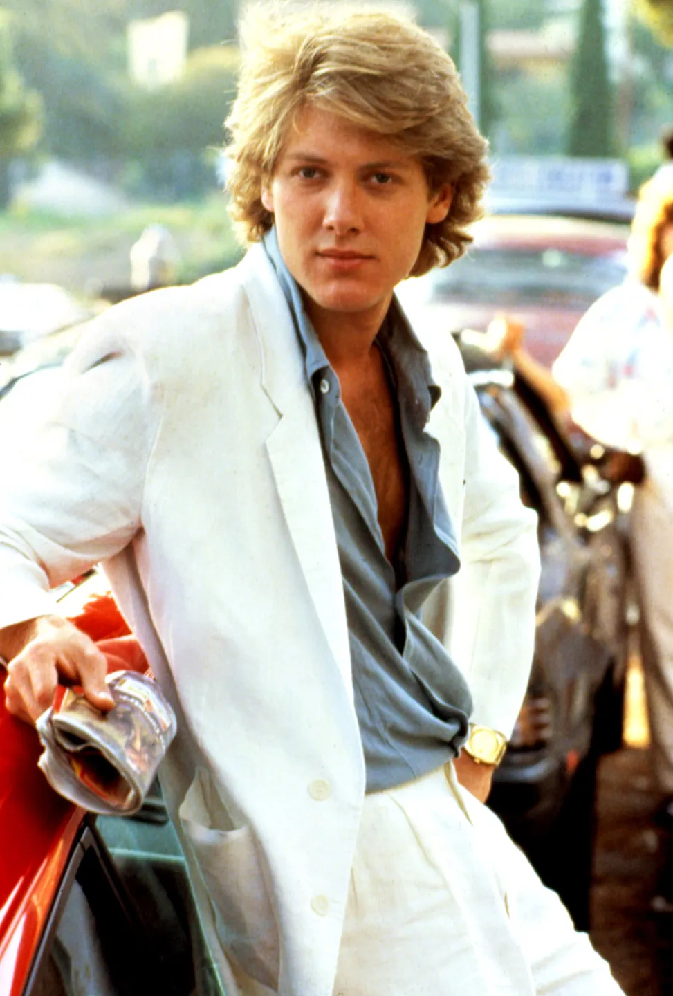 A still of a young James Spader in a white suite and a shirt open to expose his whole chest, hair feathered in true mid-1980s wealthy youth style.