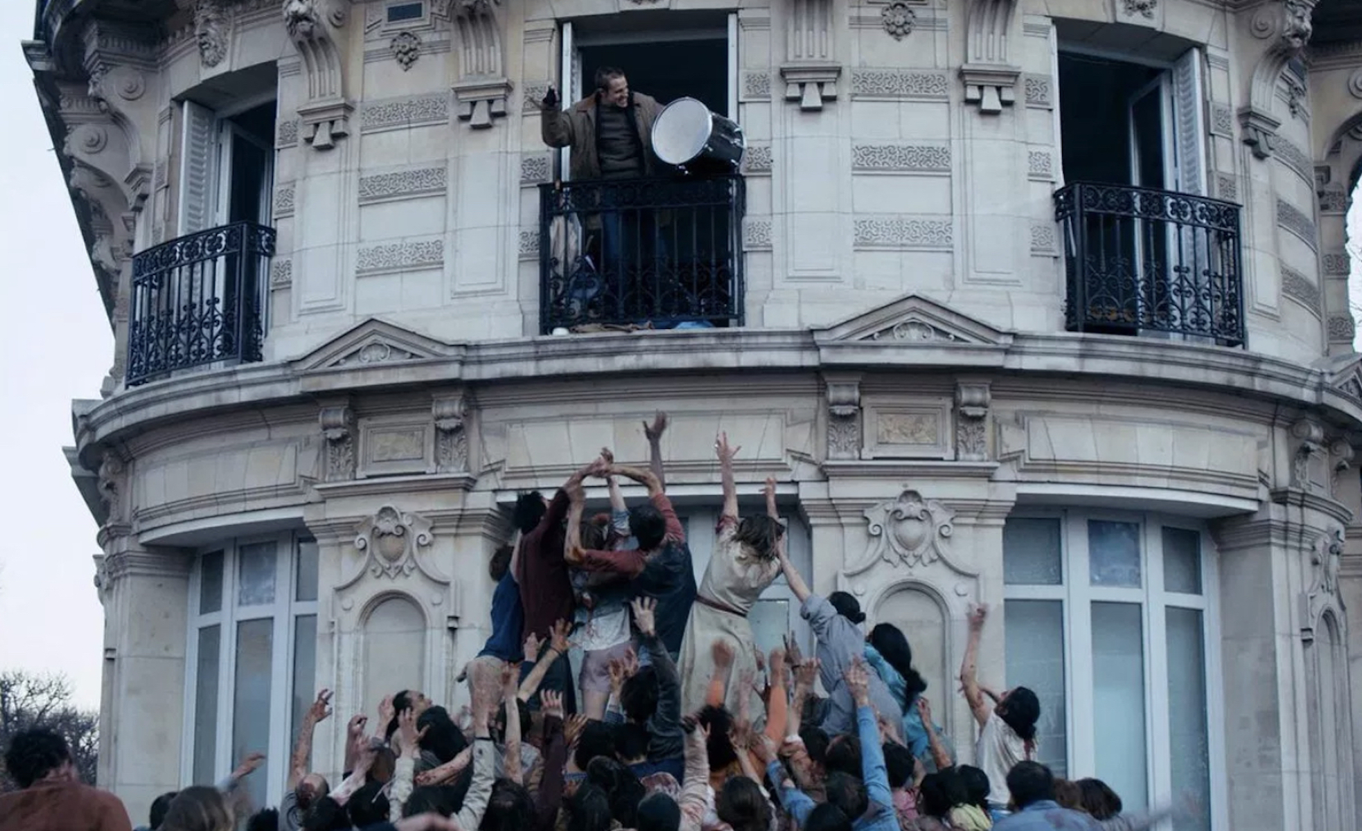 A person beats a drum out of a second floor window while hordes of zombies attempt to climb the wall to reach them.
