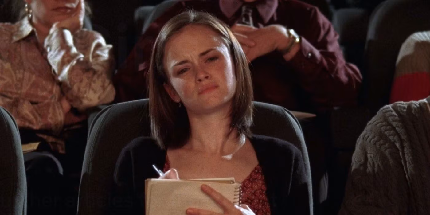 A young woman seated in an auditorium looks perplexed as she takes notes in a spiral notebook.